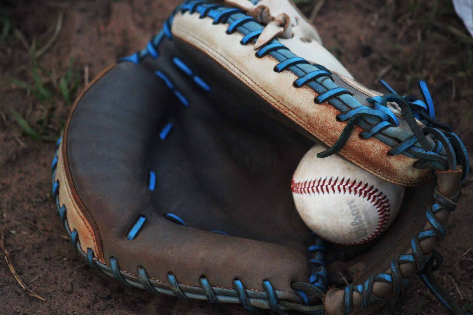 Close-up of a worn leather baseball glove with blue laces holding a baseball on infield dirt.