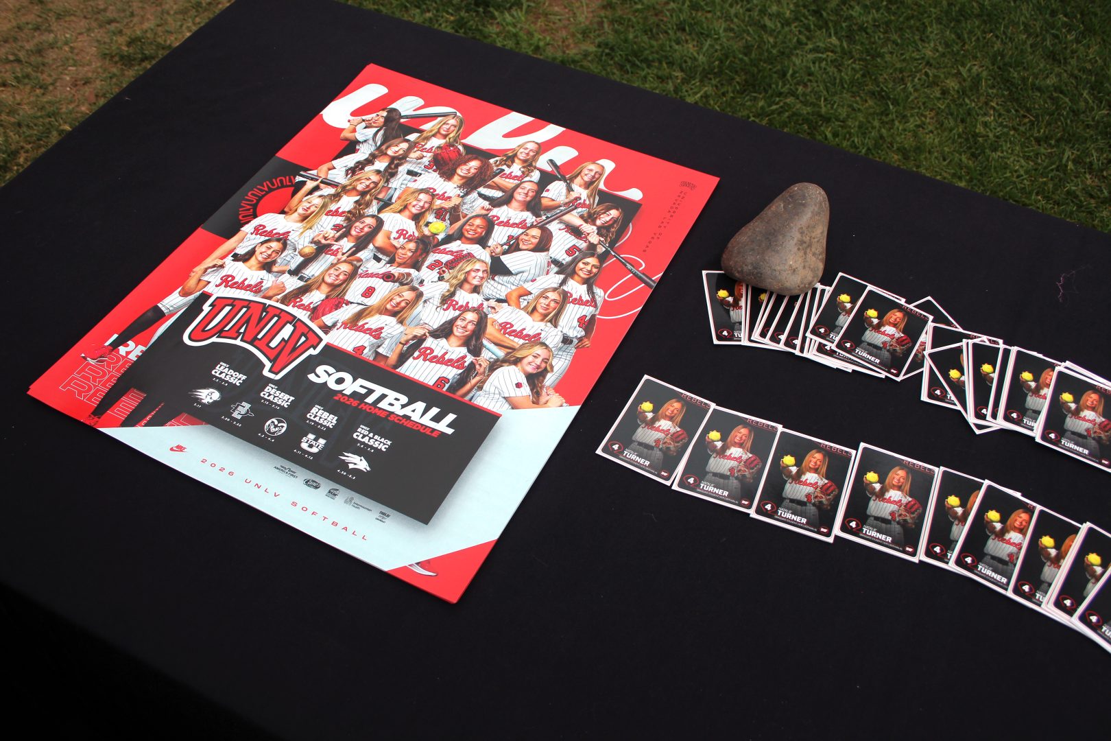 UNLV Softball 2026 poster and schedule on a black table outdoors with a stack of Natalie Turner player cards held down by a rock.