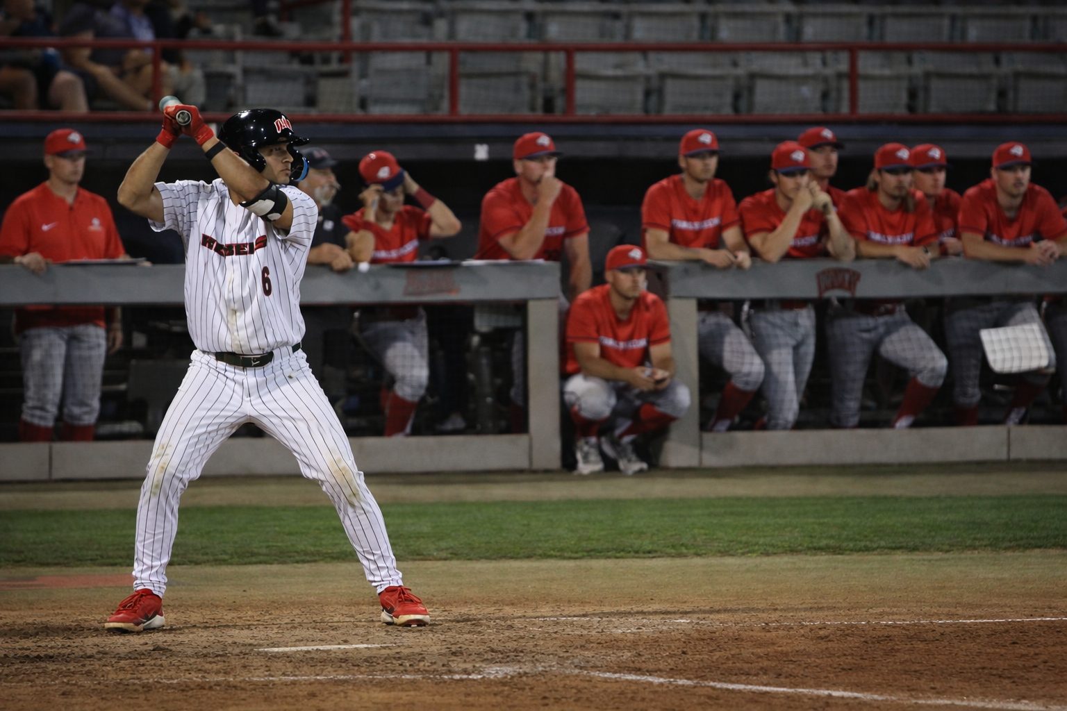 UNLV second baseman Marcos Rosales (#6) stands in the batter’s box in a white pinstriped Rebels uniform as the catcher and home-plate umpire set behind him, with the opposing dugout and fans in the background.