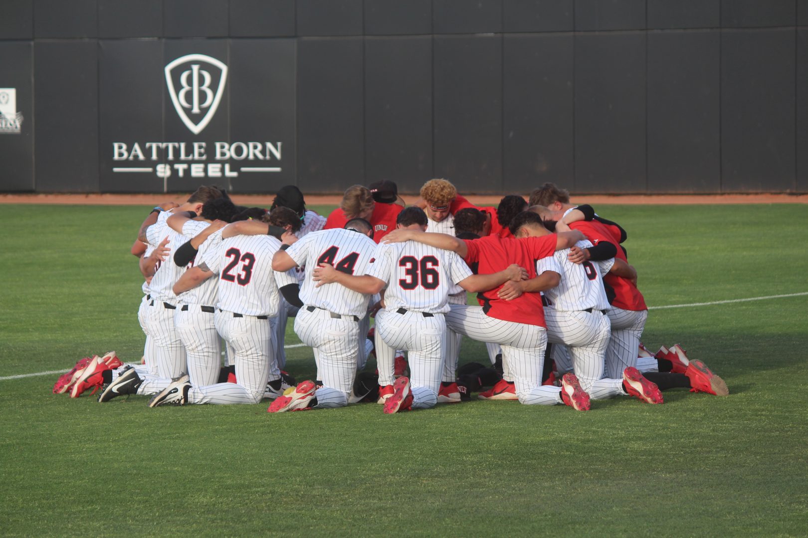 UNLV baseball players kneel in a tight pregame huddle on the outfield grass at Earl E. Wilson Stadium, arms around each other, with the “Battle Born Steel” sign on the outfield wall behind them.
