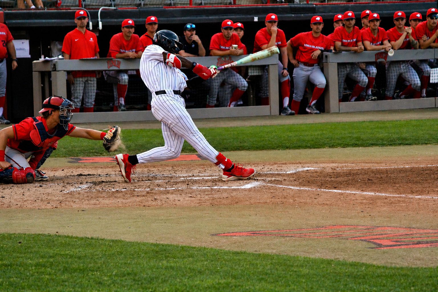 Reggie Bussey swings at a pitch during a UNLV baseball home game, with the catcher reaching for the ball and the opposing team watching from the dugout.
