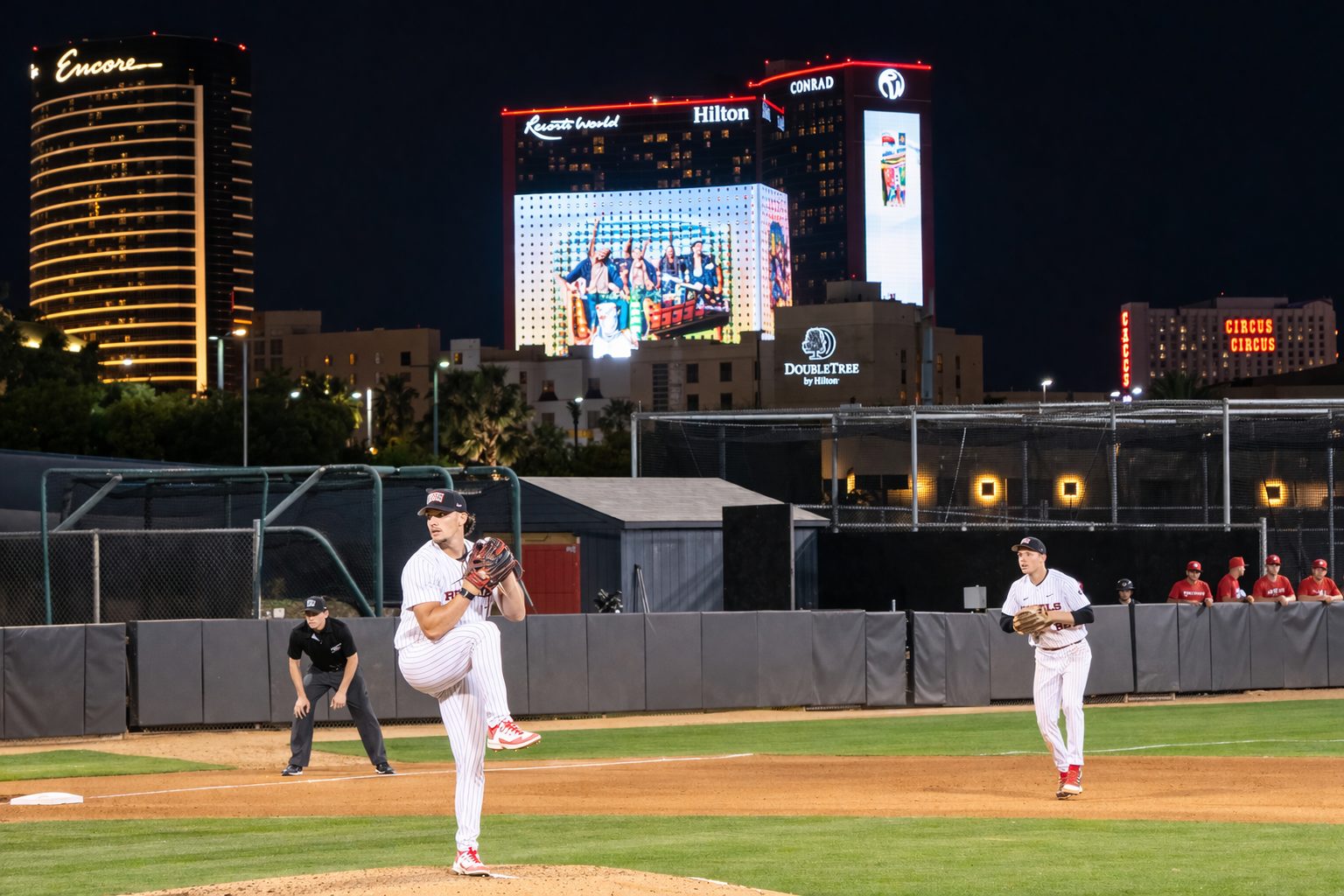 UNLV pitcher Parker Dillhoff delivers a pitch during a night game at Earl E. Wilson Stadium with the Las Vegas Strip skyline in the background.