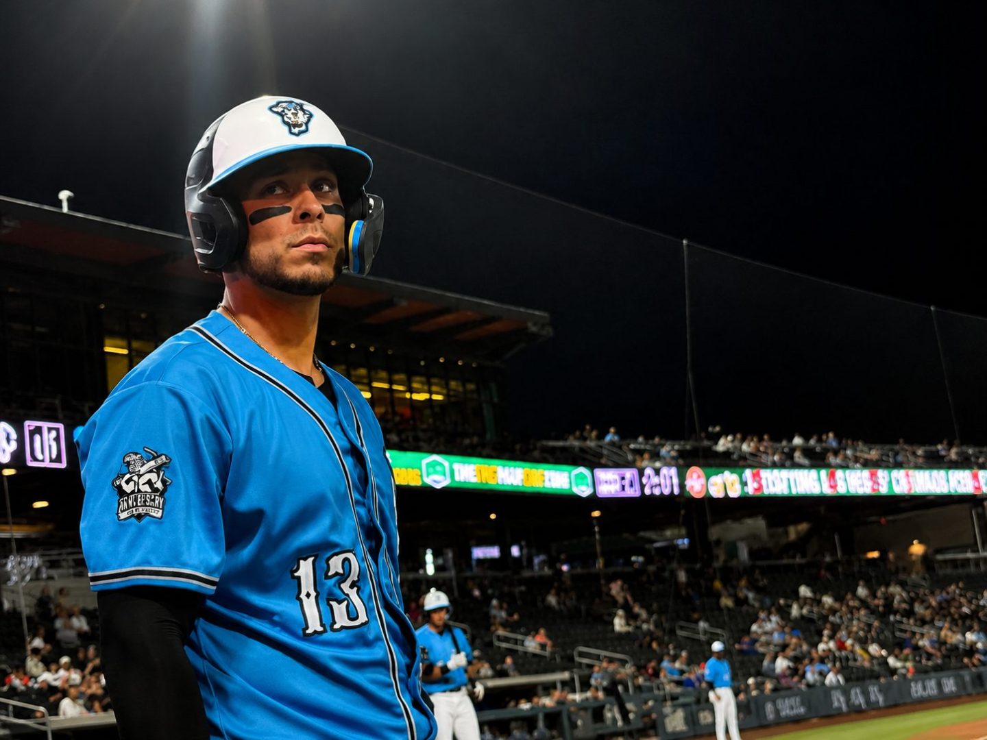 Joey Meneses wearing a blue Las Vegas Aviators jersey and batting helmet stands near the dugout during a night game at Las Vegas Ballpark.