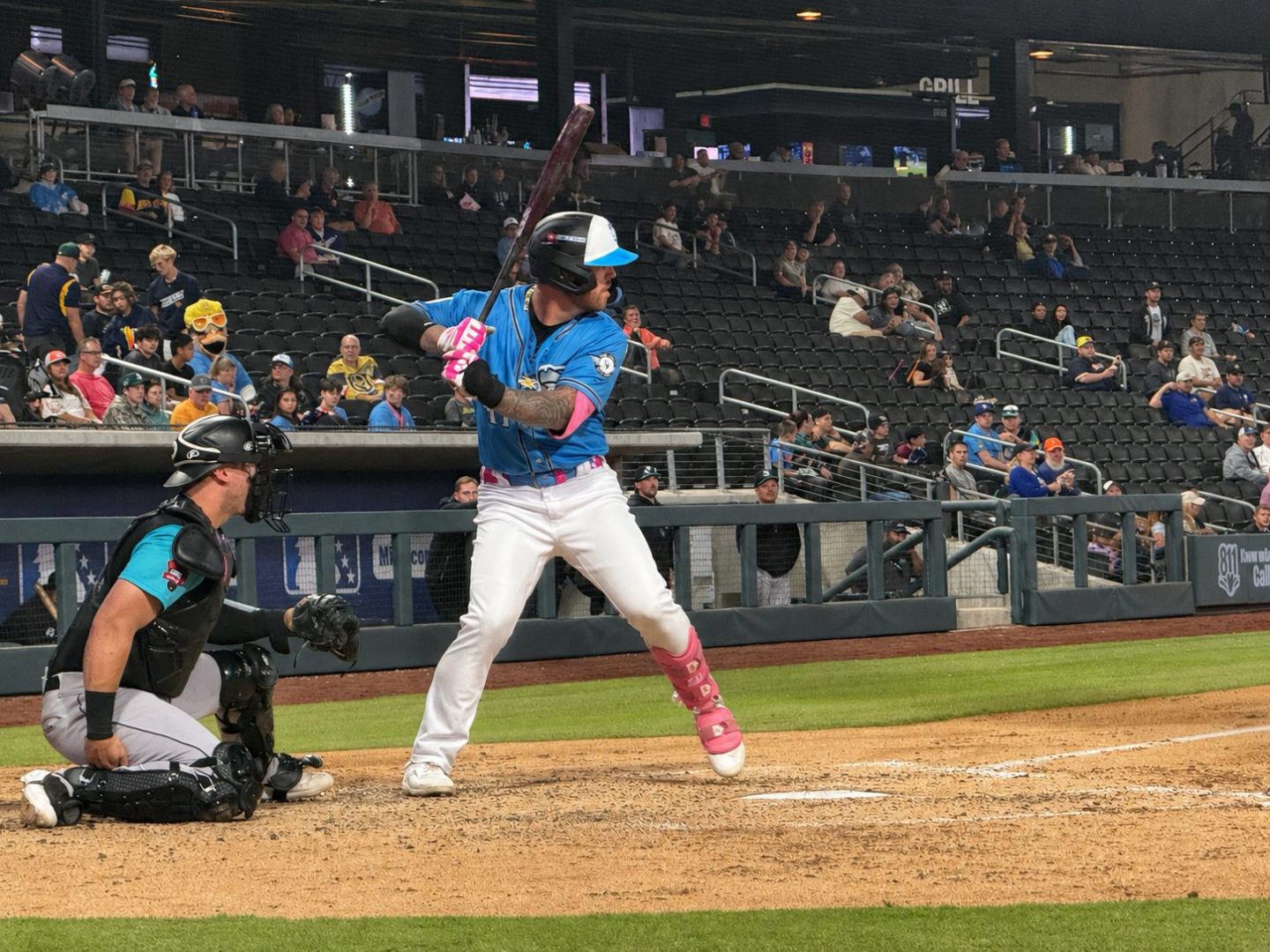 Tommy White stands in the batter’s box wearing a blue Las Vegas Aviators jersey and pink protective gear while a catcher sets up behind him during a night game.
